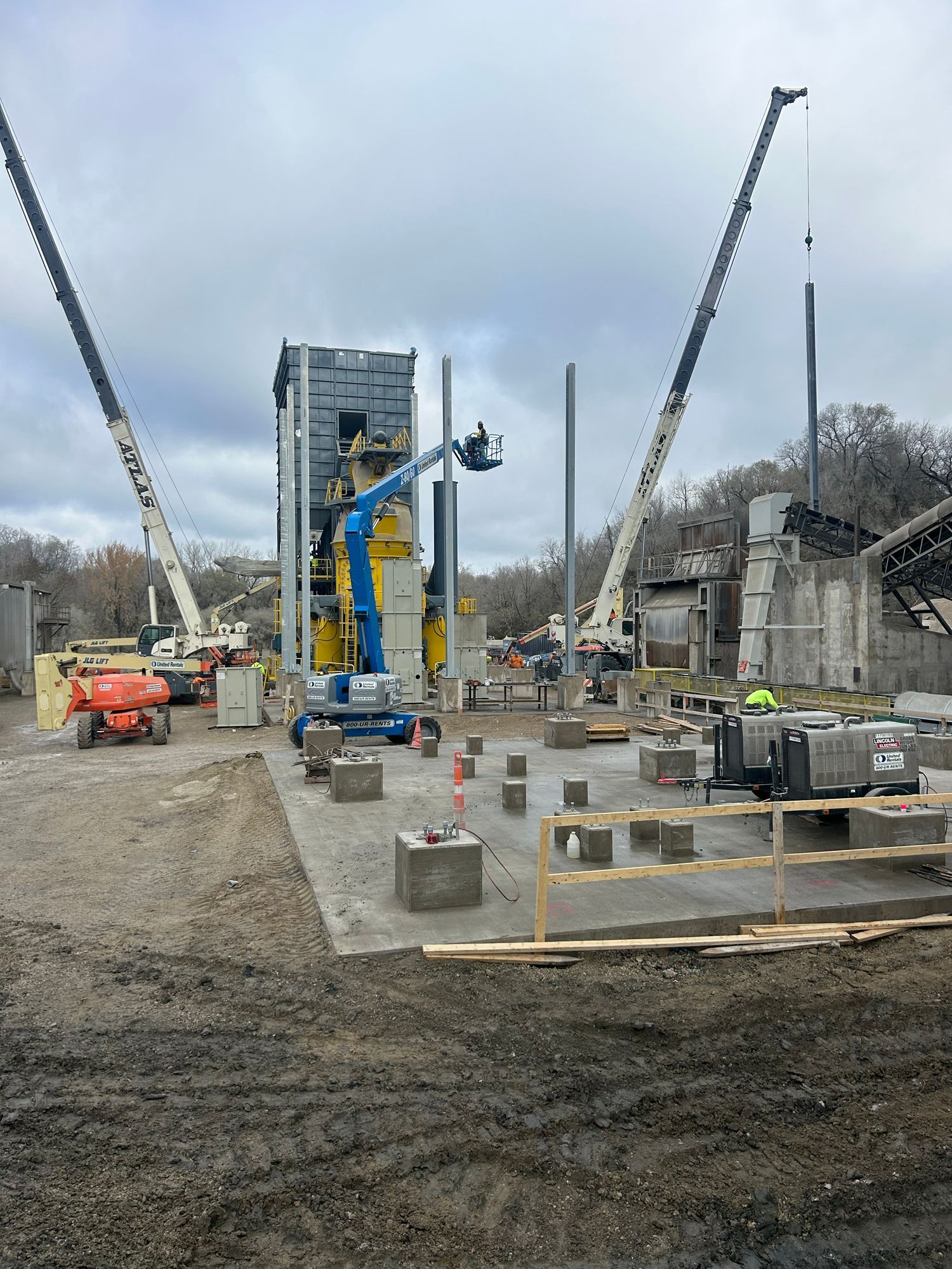 Construction site with cranes, a blue lift, and workers near a tall structure and concrete blocks.
