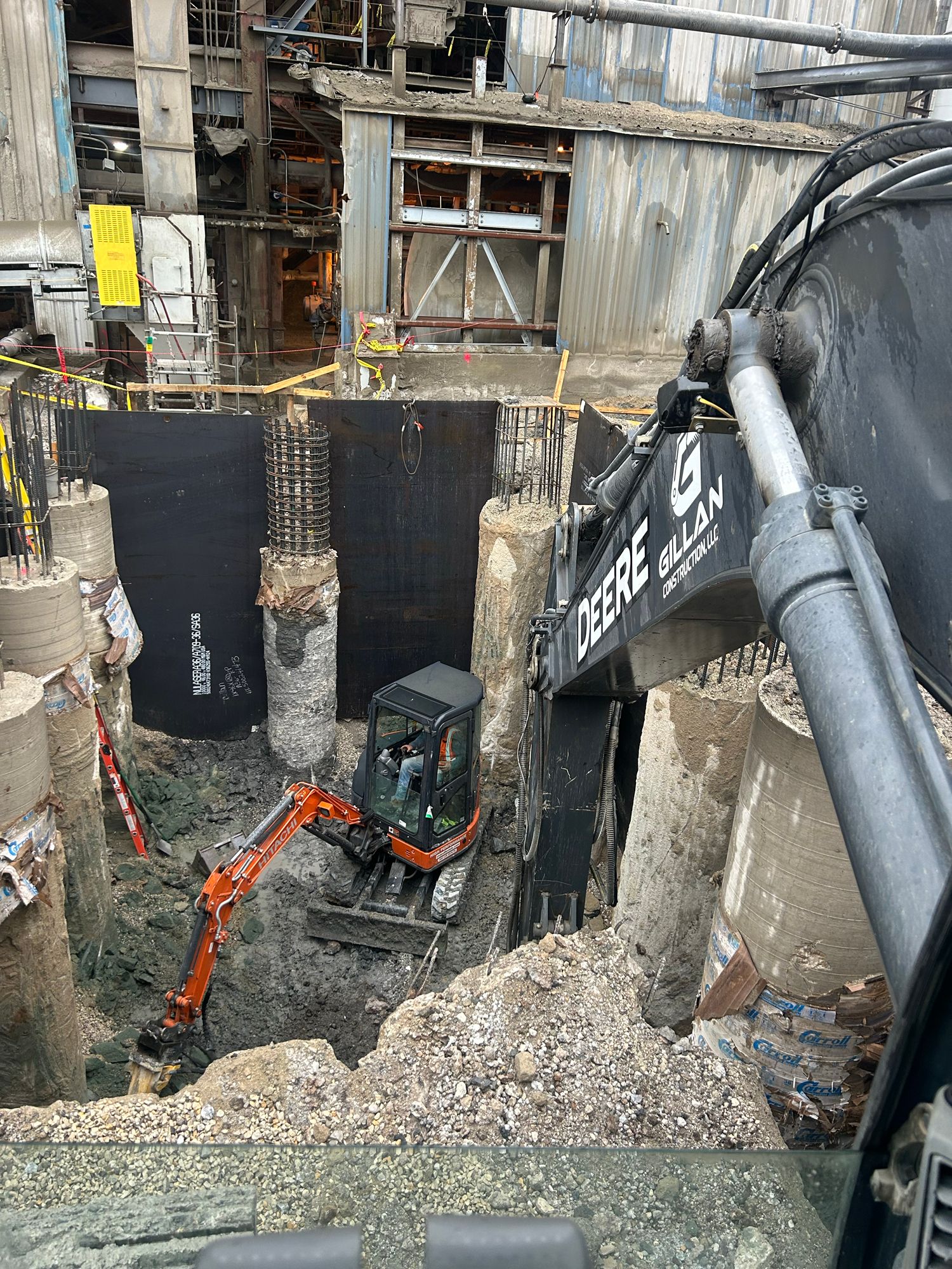 Construction site with a small excavator digging; concrete and rebar structures visible.
