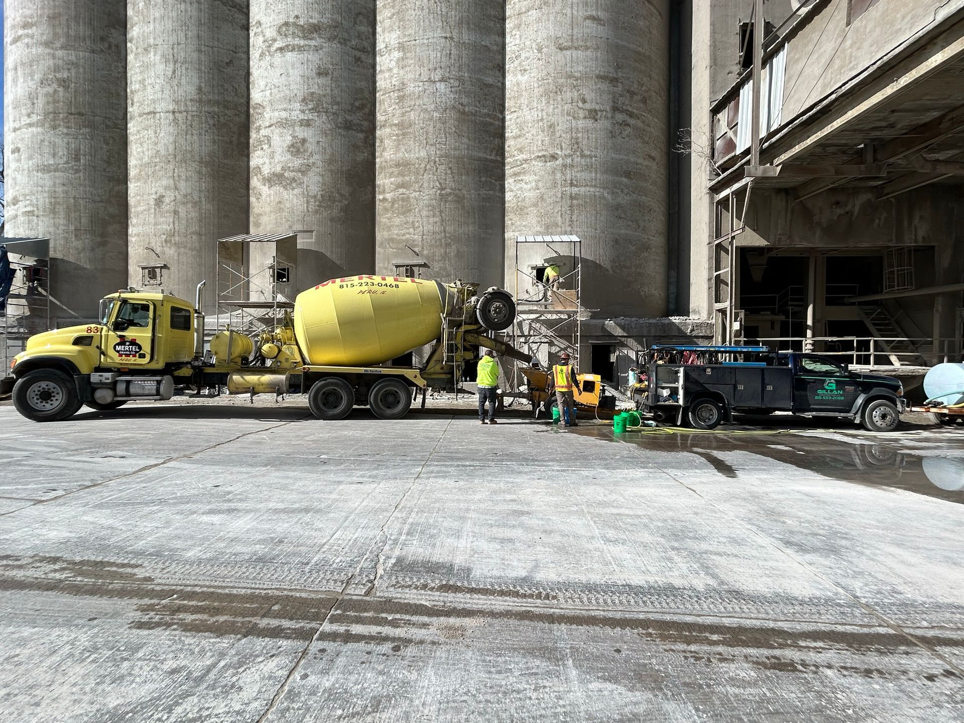 Yellow concrete truck beside a silver vehicle at a cement plant. Workers stand near them; tall cement silos are in the background.