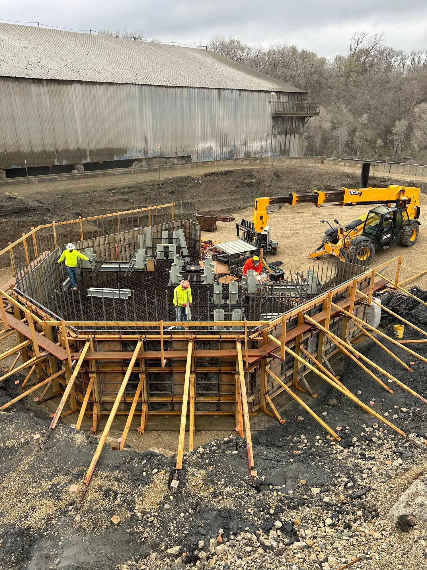 Construction site: Workers building concrete foundation; yellow machinery and a large wooden structure in the background.
