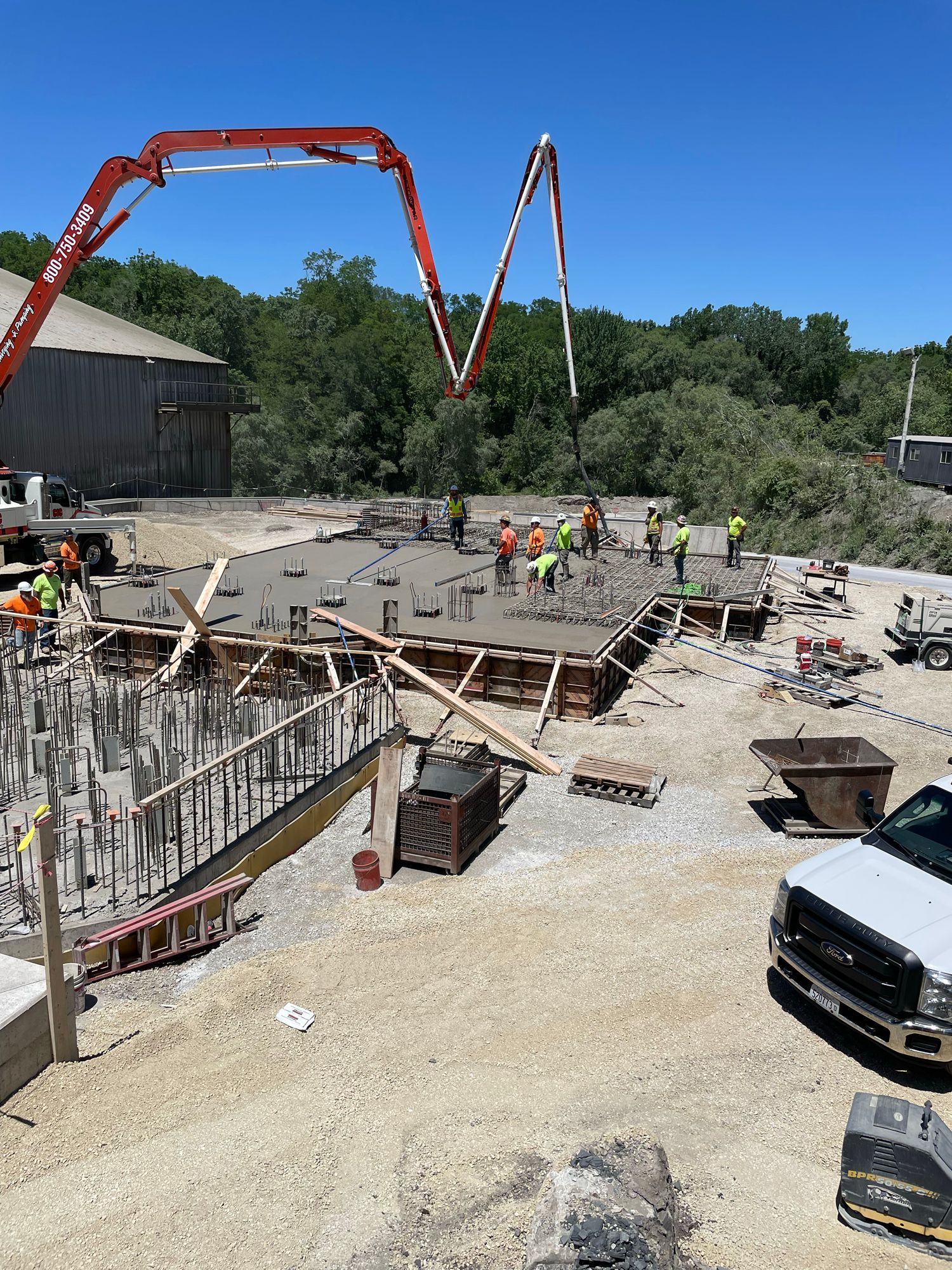 Construction site: concrete being poured onto a foundation. Workers in safety vests, red concrete pump truck. Sunny day.