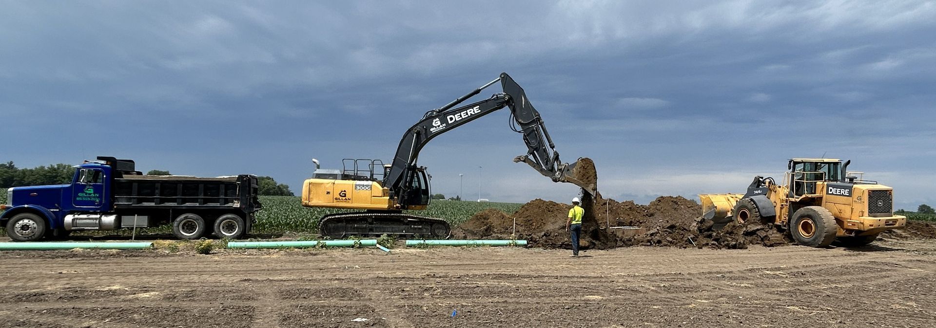 Construction scene: Excavator digging, dump truck, and front-end loader on dirt field under a cloudy sky.