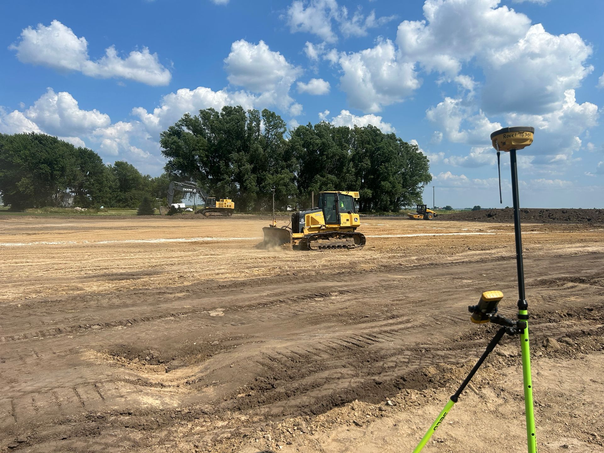Construction site with heavy machinery grading the land; GPS equipment in foreground under a blue sky.