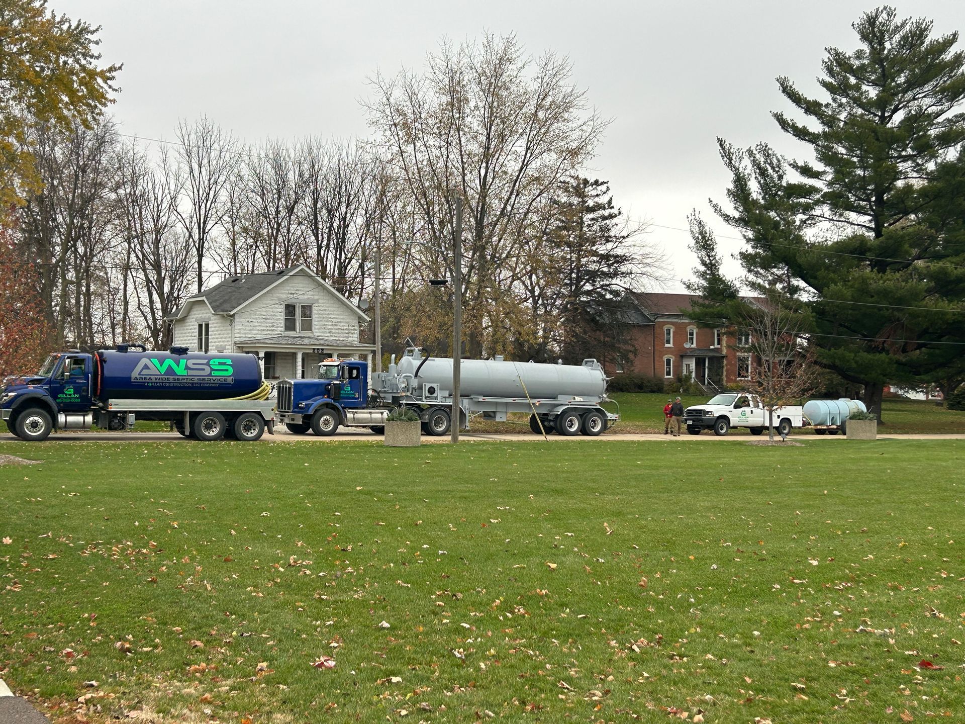 Four tanker trucks lined up on grass in front of houses; cloudy day.