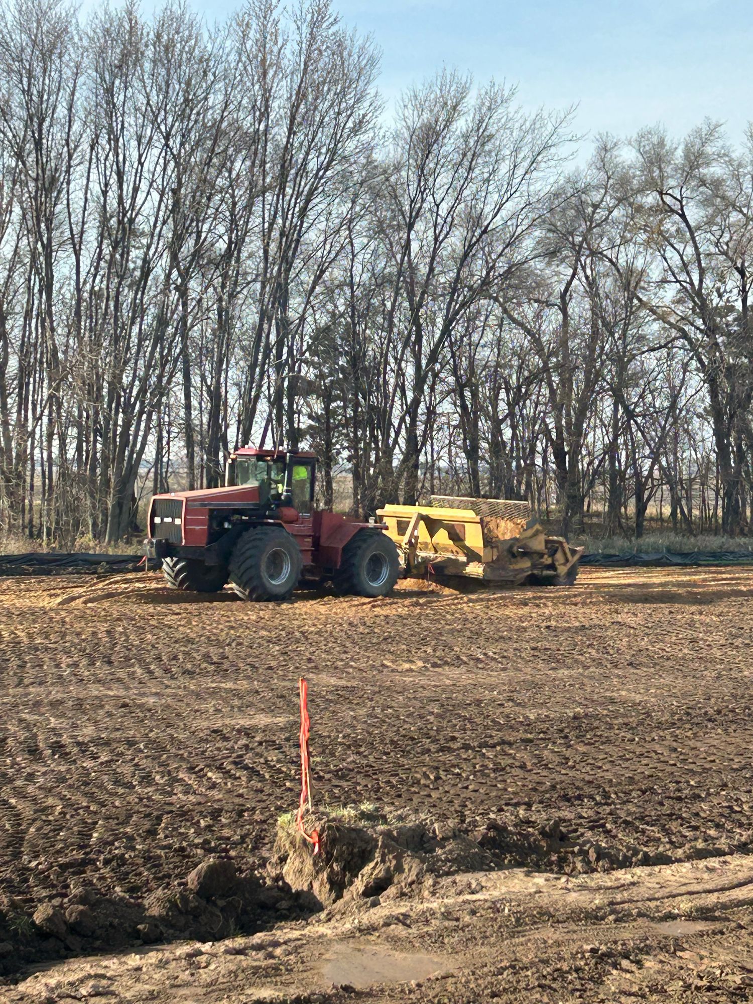 Red tractor pulling a yellow scraper on a dirt field, trees in the background.