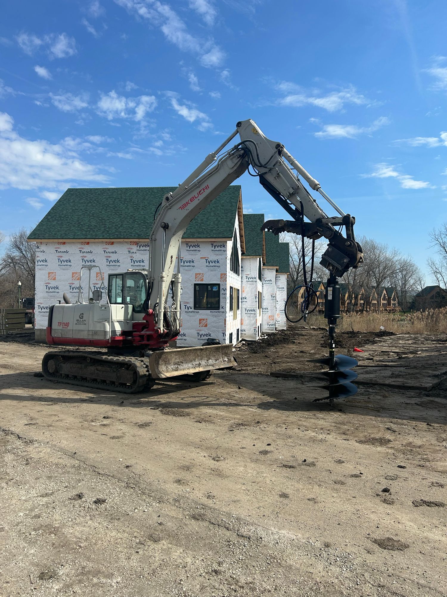 Excavator with auger drilling into the ground, construction site with unfinished houses in the background.