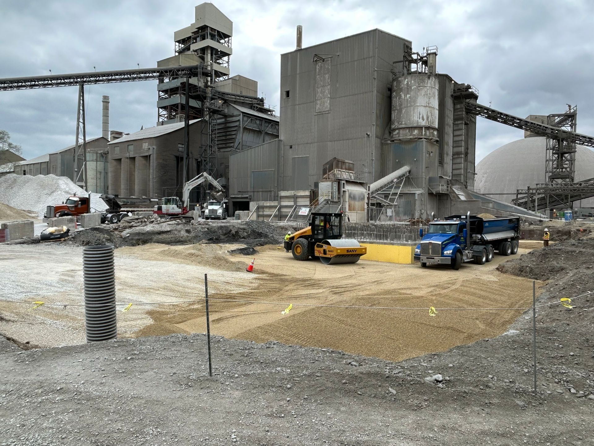 Construction site in front of a large industrial building, with machinery and gravel. Cloudy sky.