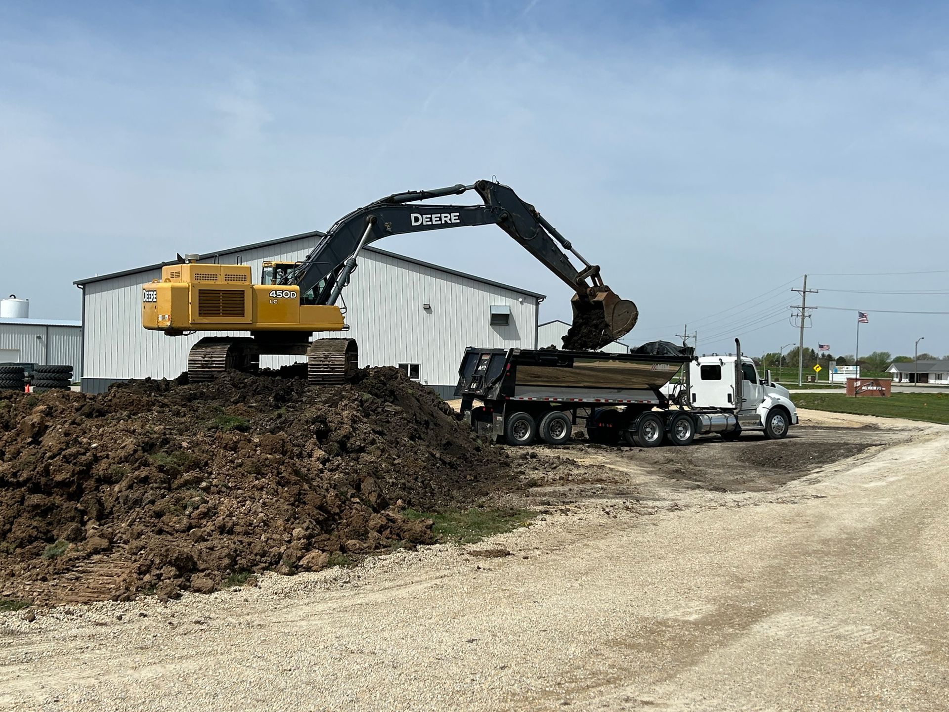 Excavator loading dirt into a dump truck, parked near a white building, on a gravel lot.