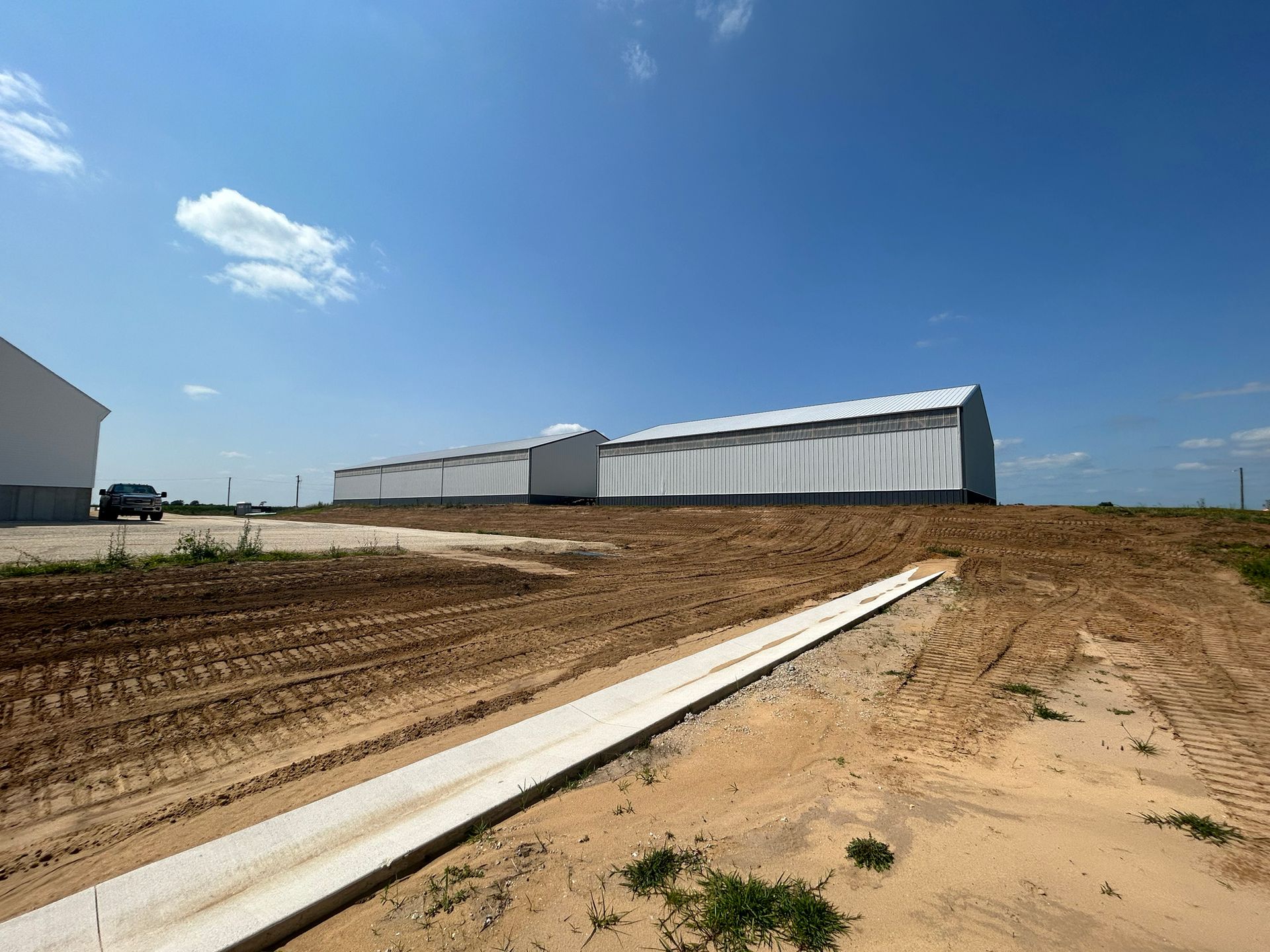 Several large, white agricultural buildings on a dirt lot under a bright blue sky.