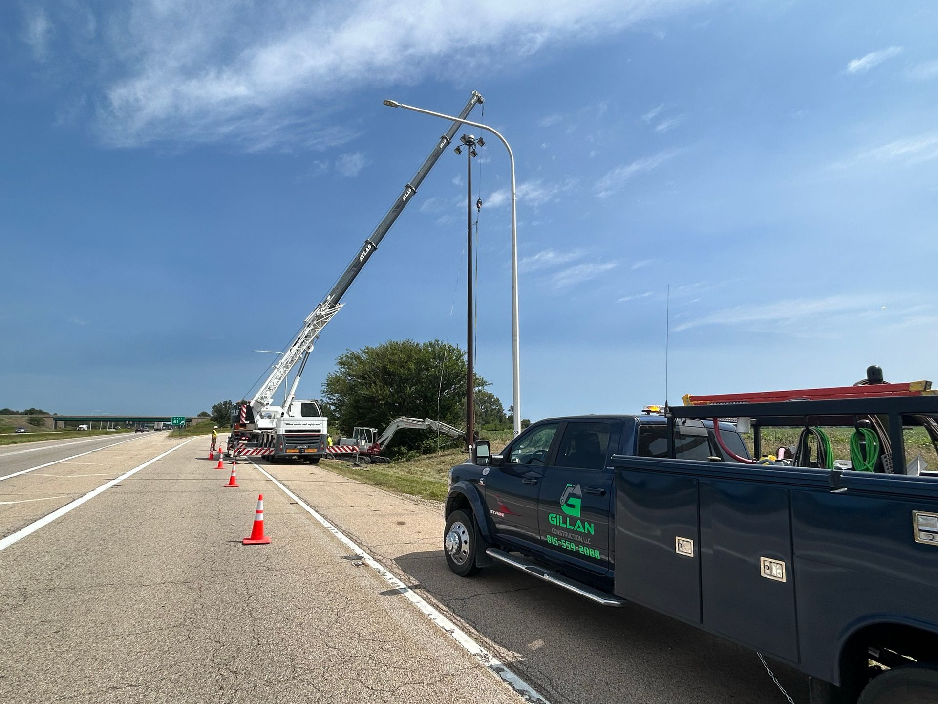 A crane lifts a street light, with a service truck on a highway shoulder under a blue sky.