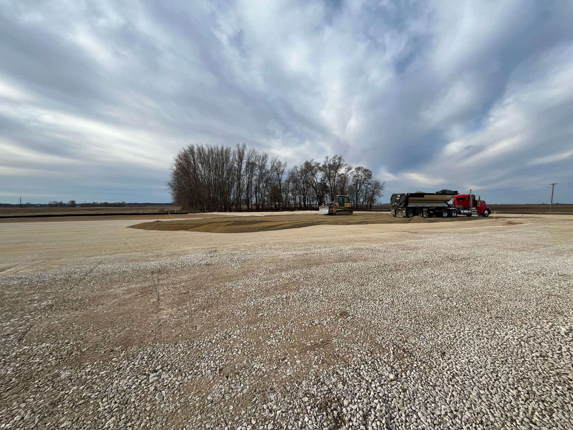 A gravel lot with a red truck, trees, and a cloudy sky.