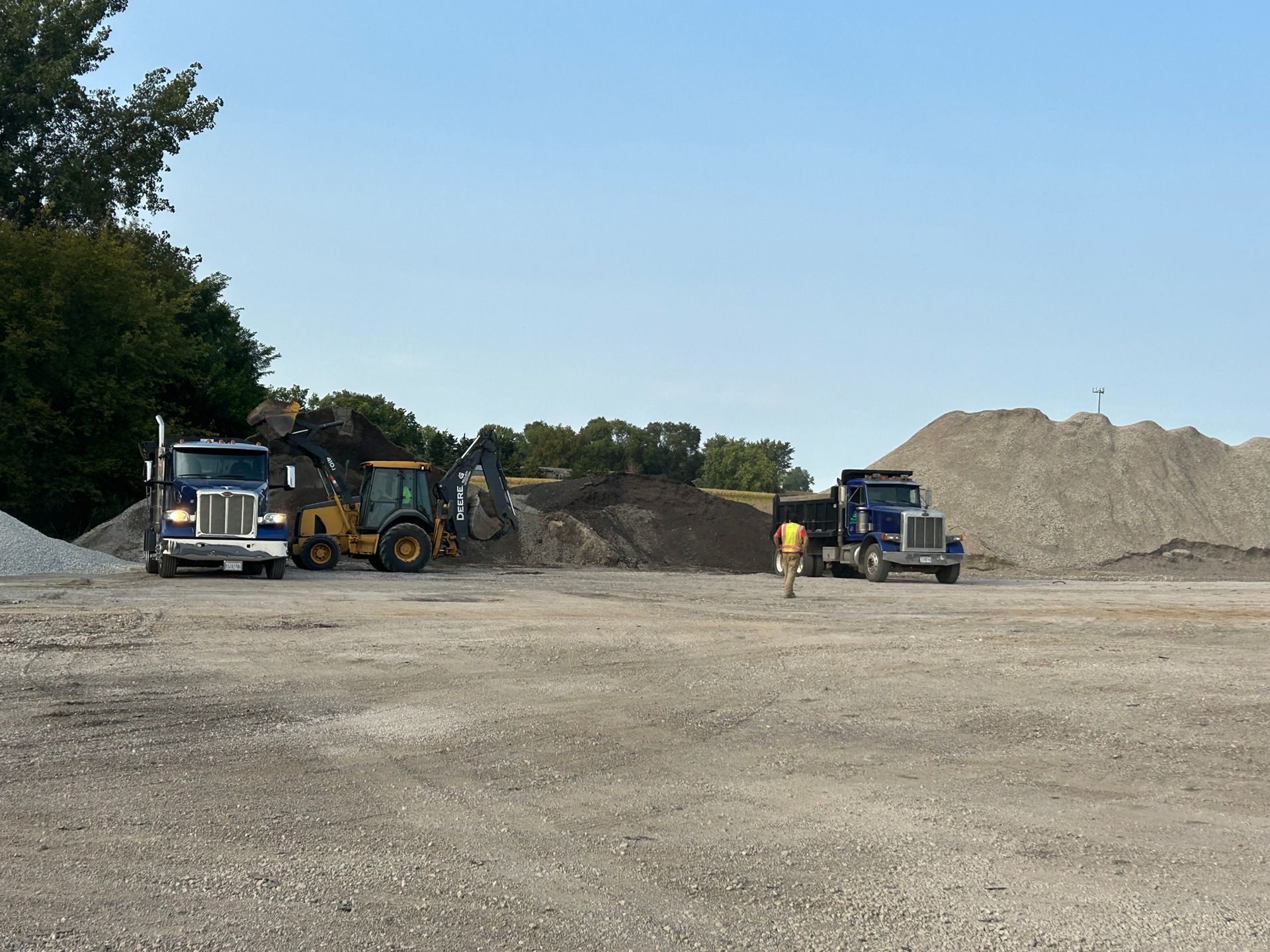 Two semi-trucks, a yellow loader, and piles of gravel at a construction site under a blue sky.