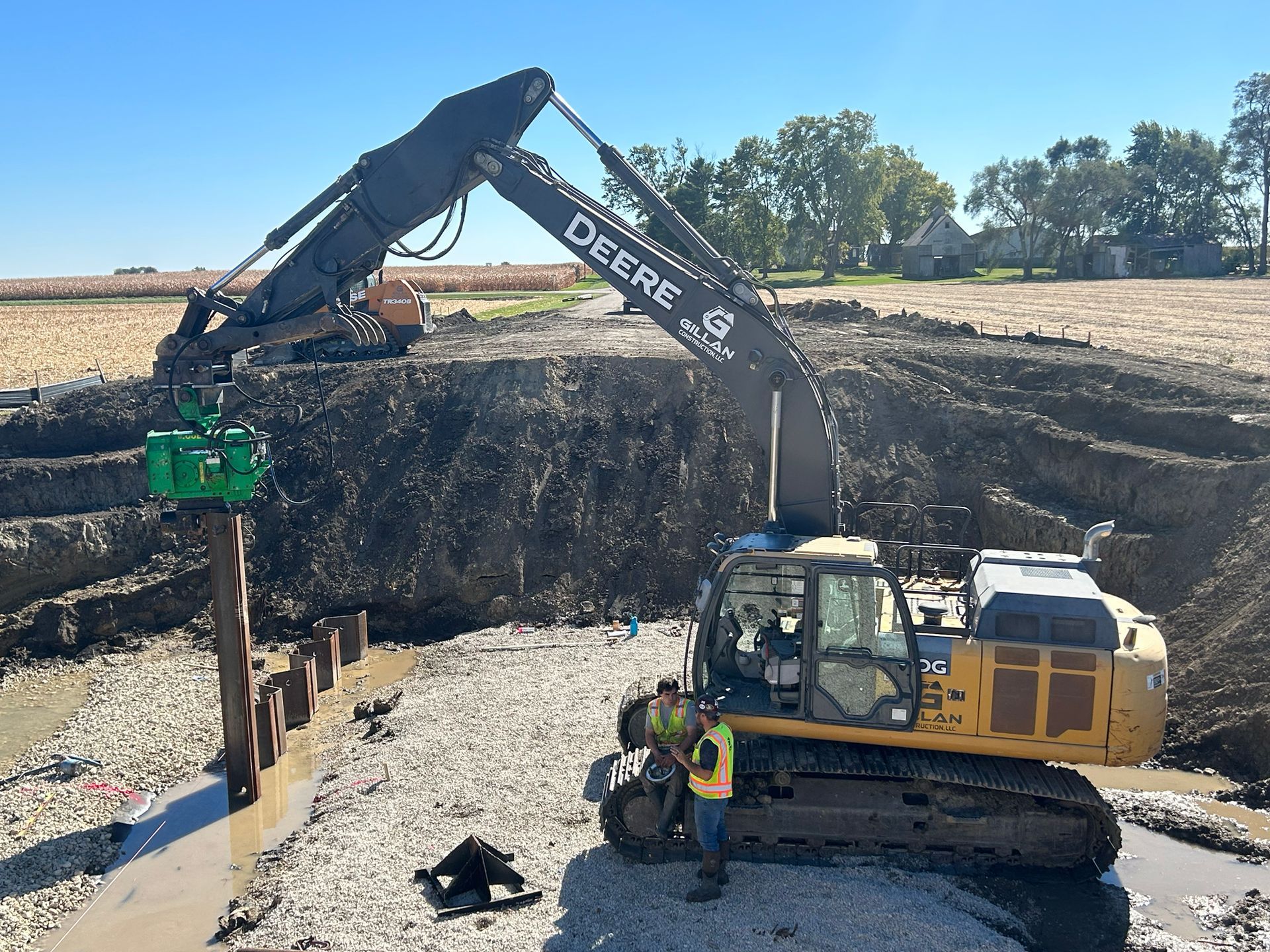 John Deere excavator placing a green device over a brown post; construction site with workers.