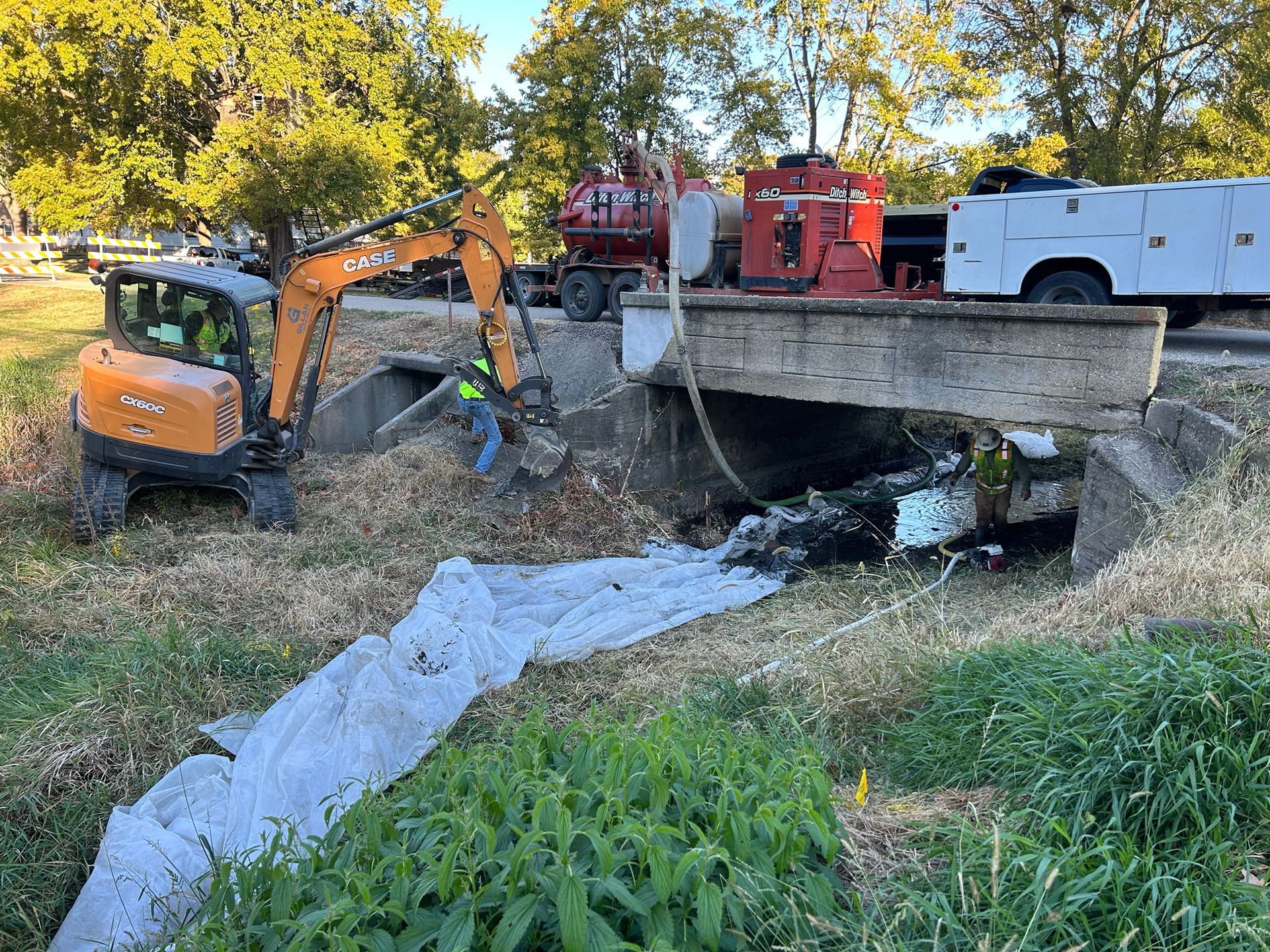 An excavator clearing debris from under a bridge.  Various trucks parked nearby. Green grass and foliage surrounding the area.
