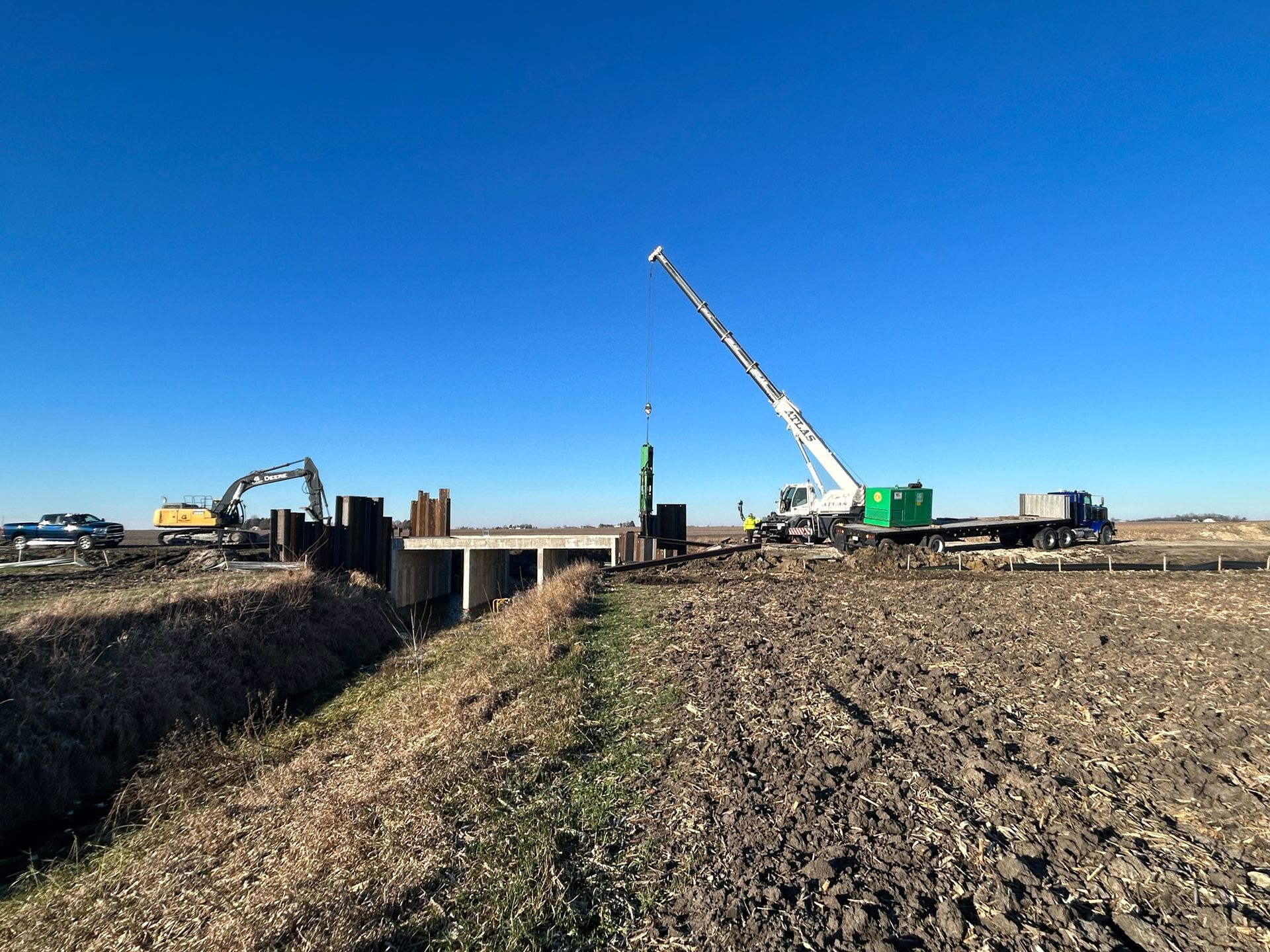 Construction site: Crane lifting a steel piling near a bridge. An excavator and truck are also visible. Bright blue sky.