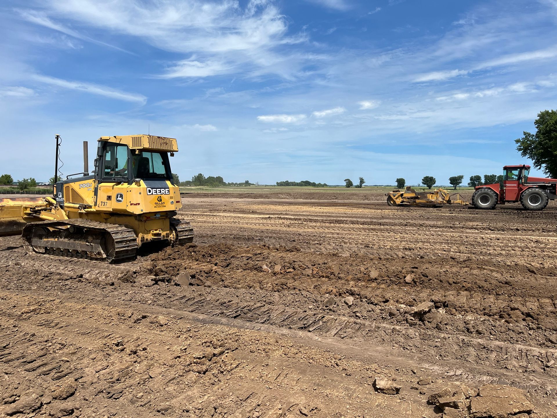 Yellow bulldozer and red tractor working in a muddy field under a blue sky.