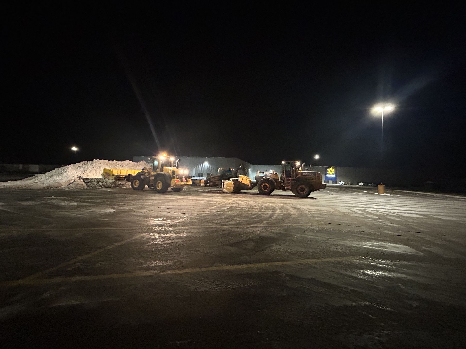 Night scene of snow removal. Several loaders are clearing a large snow pile in a parking lot. Lights illuminate the scene.