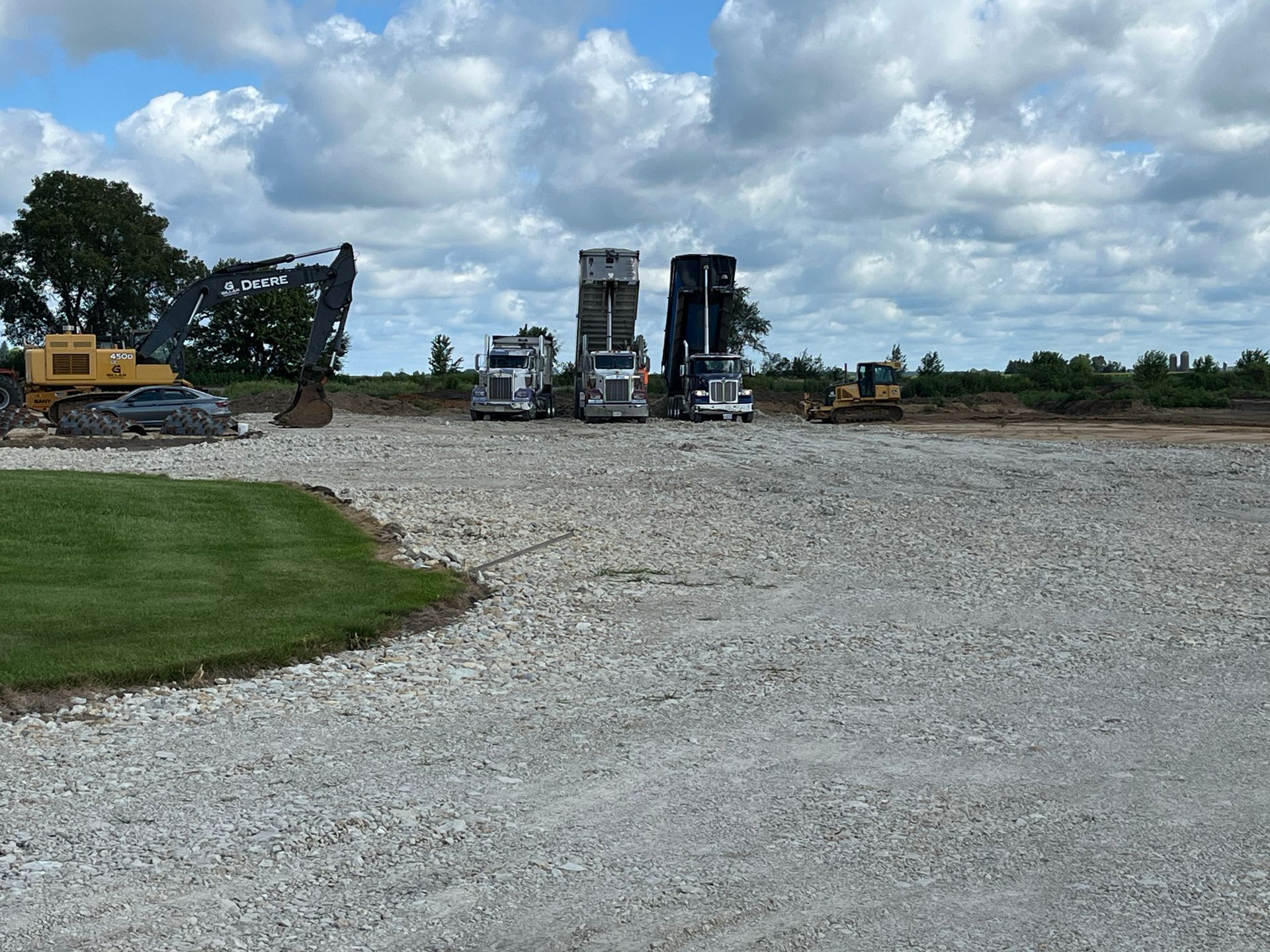 Construction site with trucks, excavators, and gravel. Cloudy sky over a partially paved area.