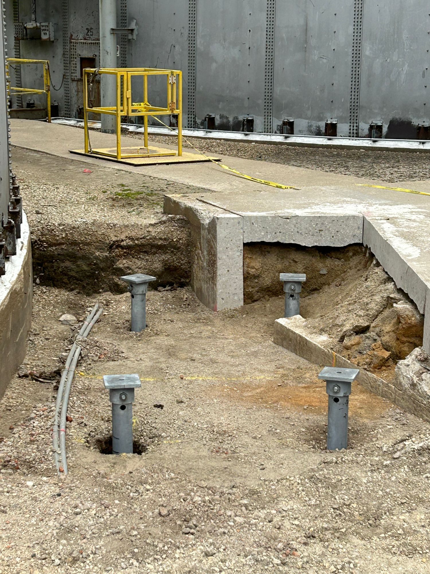Construction site with gray pipes, dirt, concrete, and yellow safety barriers near large tanks.
