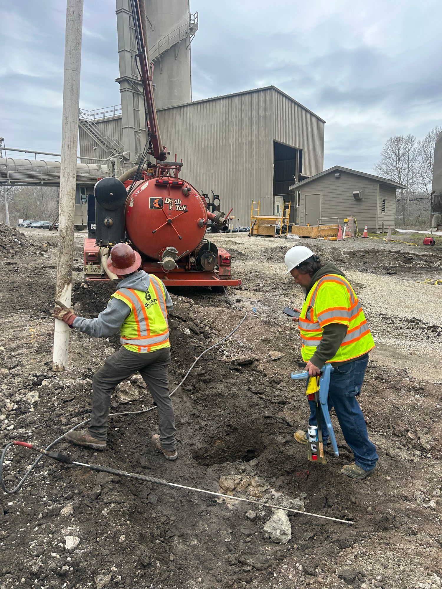Two workers in safety vests dig near red vacuum truck, utility pole, and industrial building.