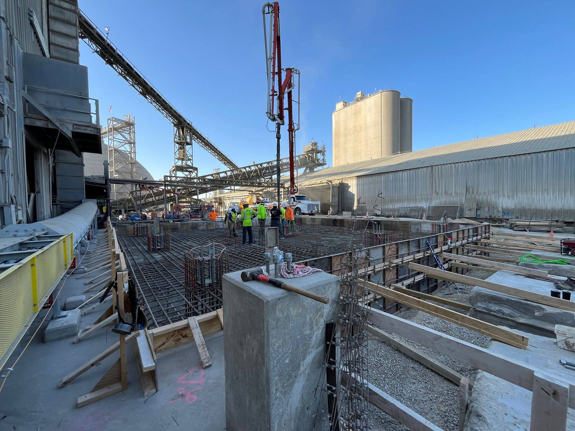 Construction site with workers, rebar, concrete forms, and a concrete pump against a clear sky.
