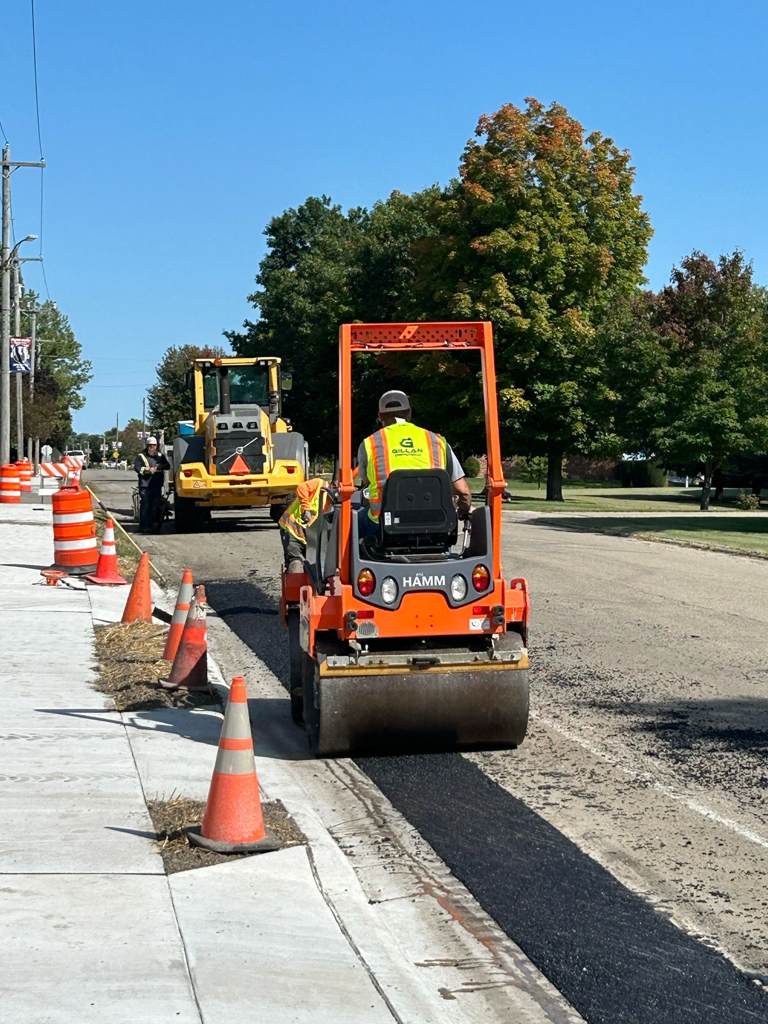 Road construction: Roller compacting asphalt strip next to sidewalk; another machine in background. Bright, sunny day.