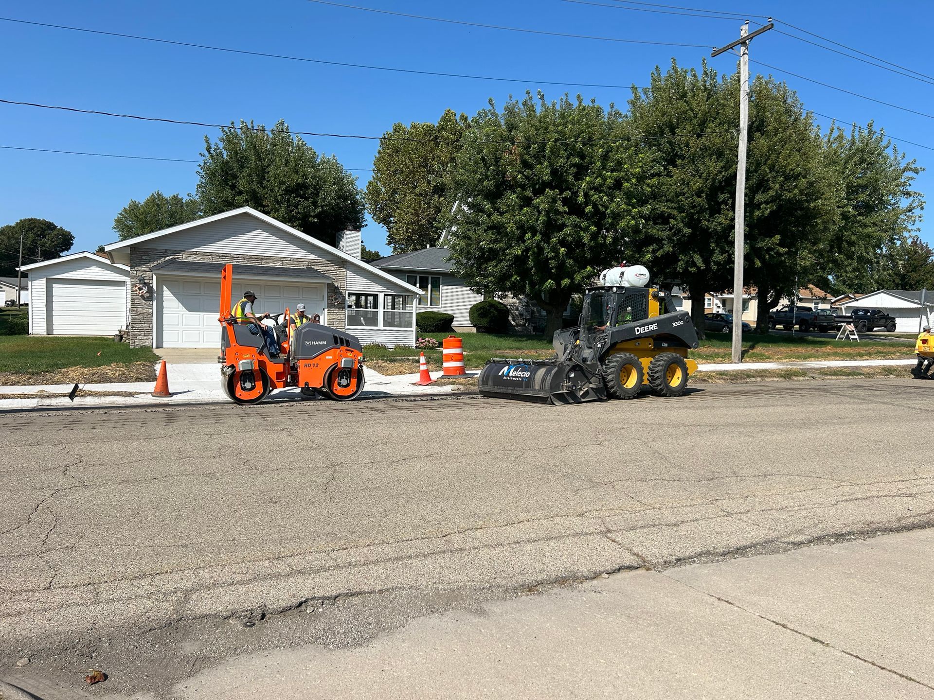Road construction: Compacted gravel road with a small roller and a front-end loader; residential setting.