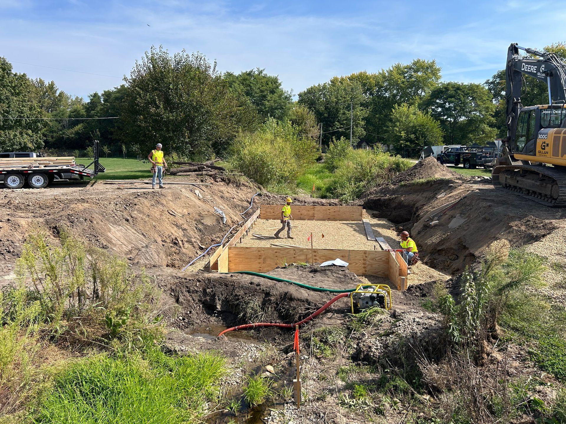 Construction site. Workers building a rectangular structure in a dirt trench, surrounded by trees and equipment.