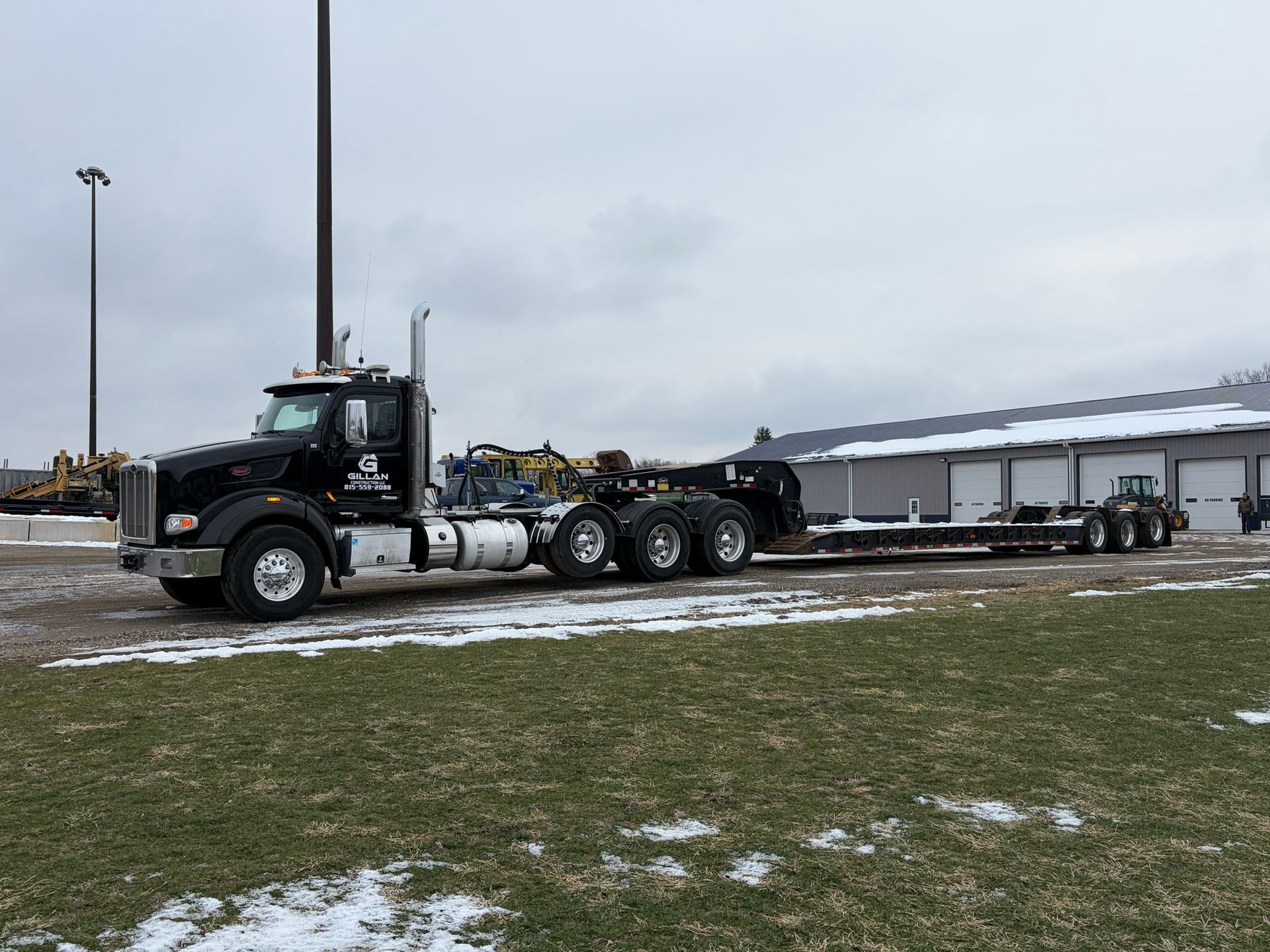 Black semi-truck pulling a lowboy trailer parked on a snowy lot near a building.