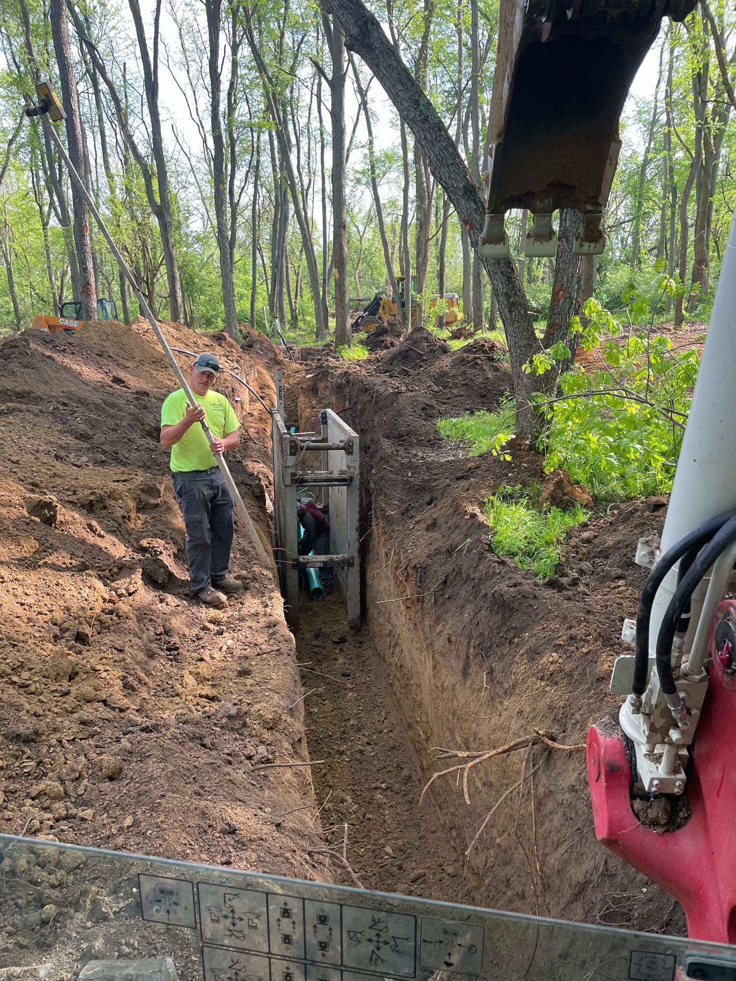 Man holding pole, assisting in excavation of a trench, two workers in trench.