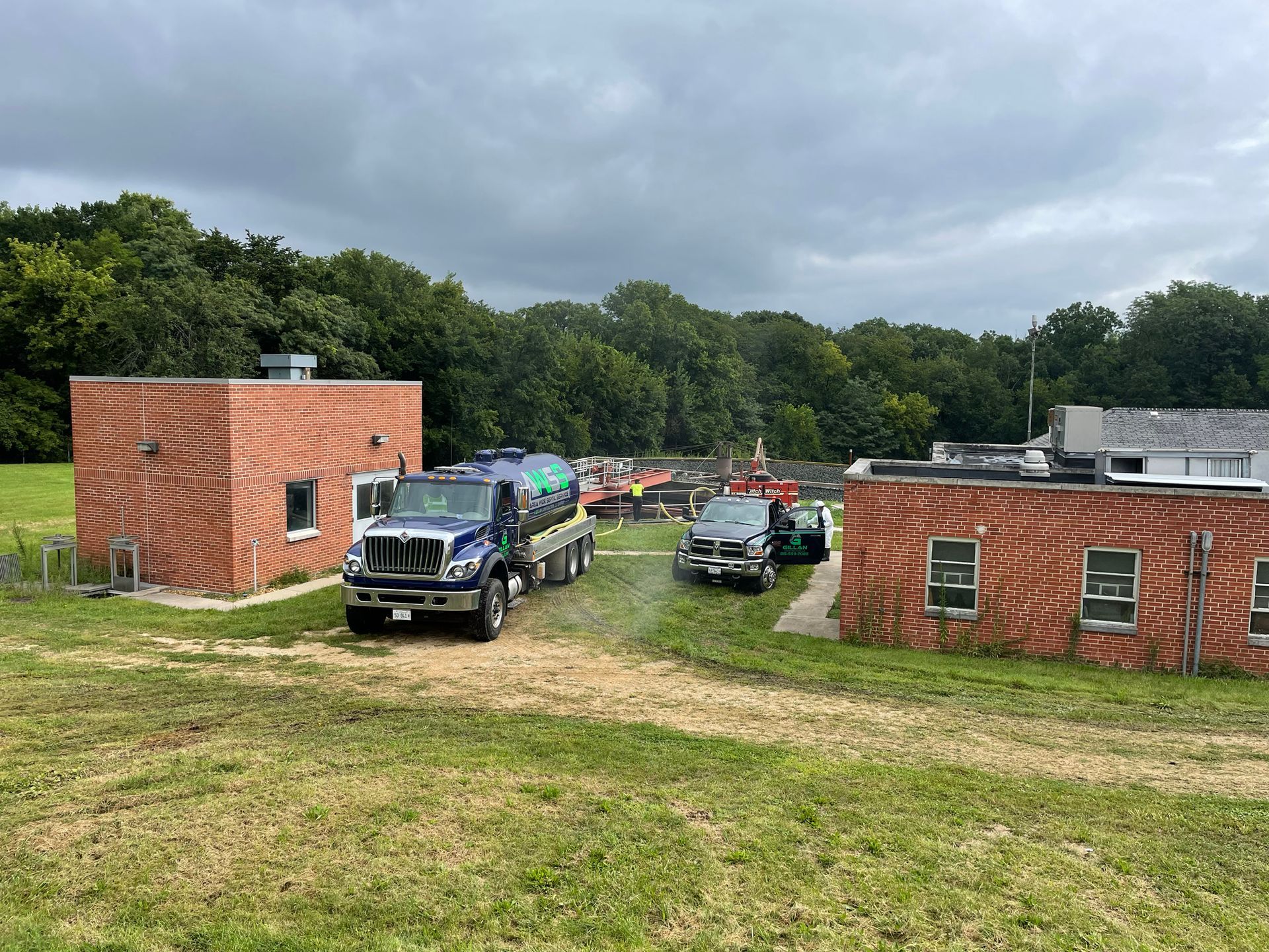 Trucks parked near brick buildings on a grassy area, trees in the background under a cloudy sky.