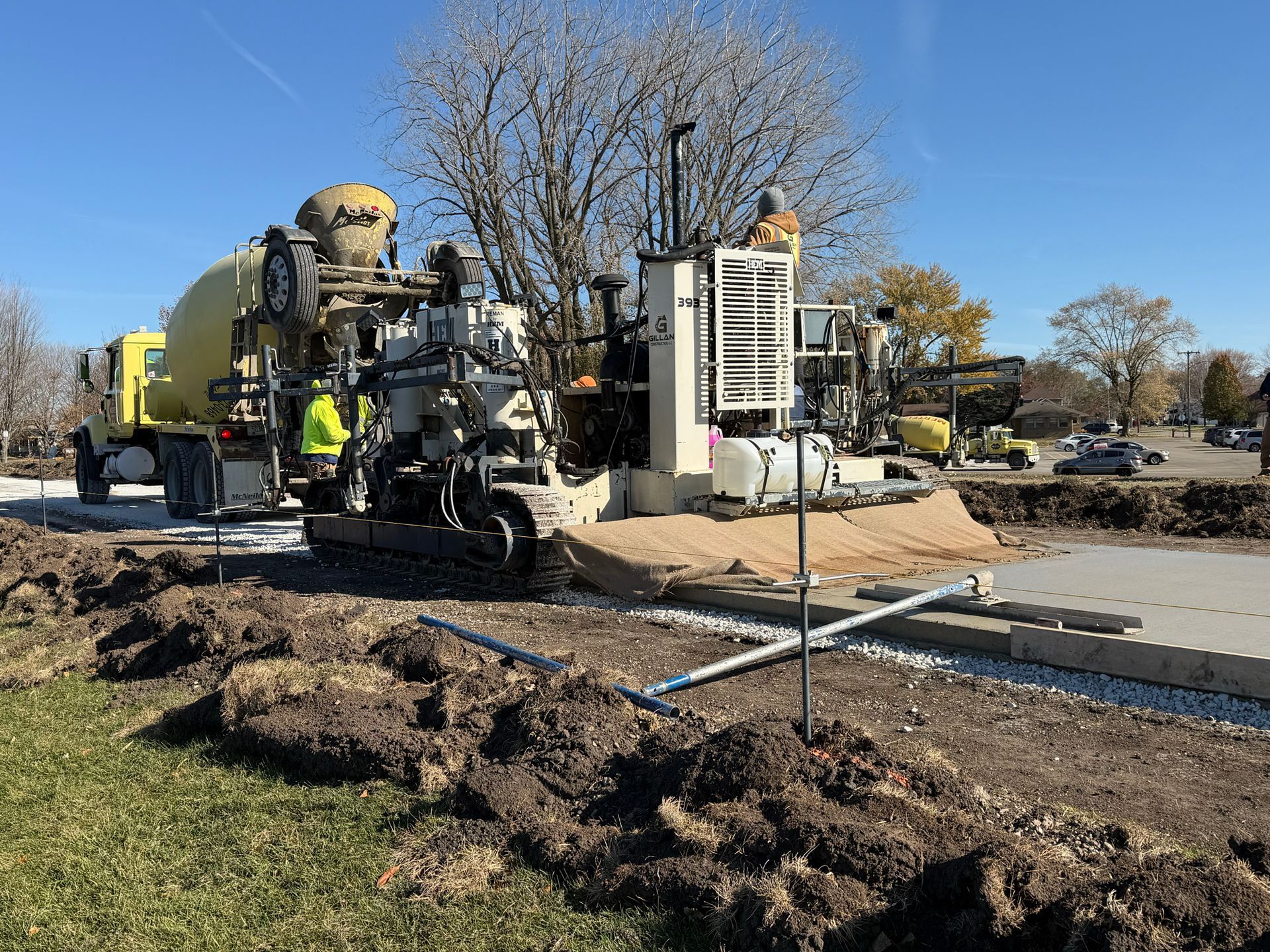 Concrete being poured by a machine at a construction site with a concrete truck, workers, and a tree.