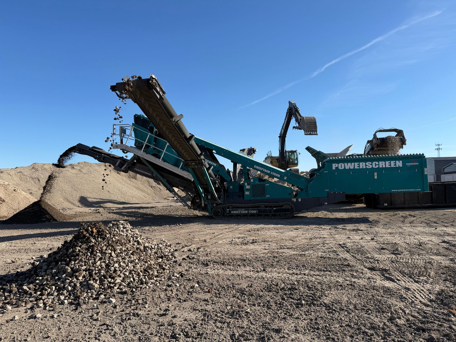 A large, green, mobile rock crusher with conveyor belts processing gravel, in an outdoor setting.