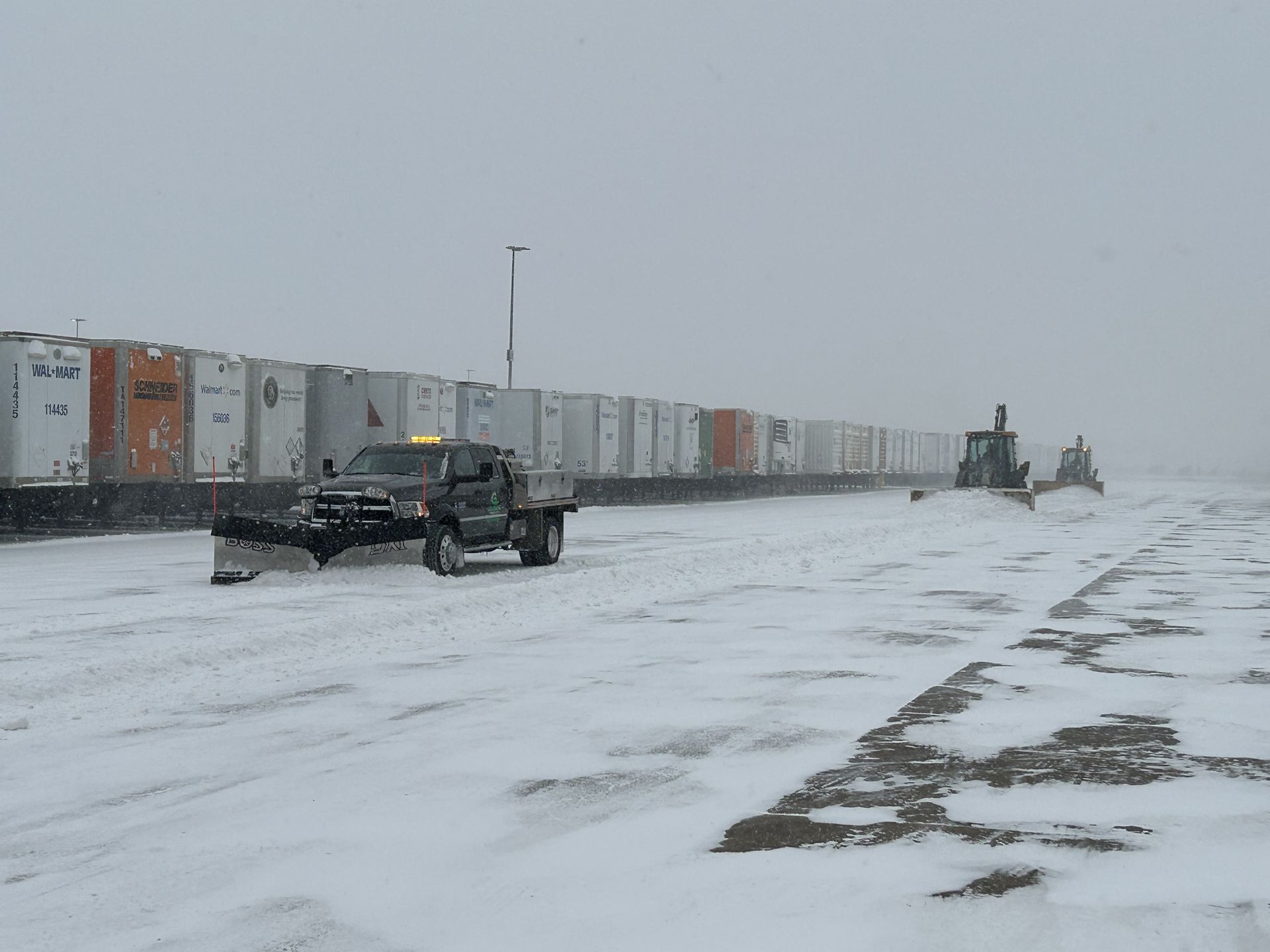 Snowplows clearing a snowy area next to a row of white containers on an overcast day.