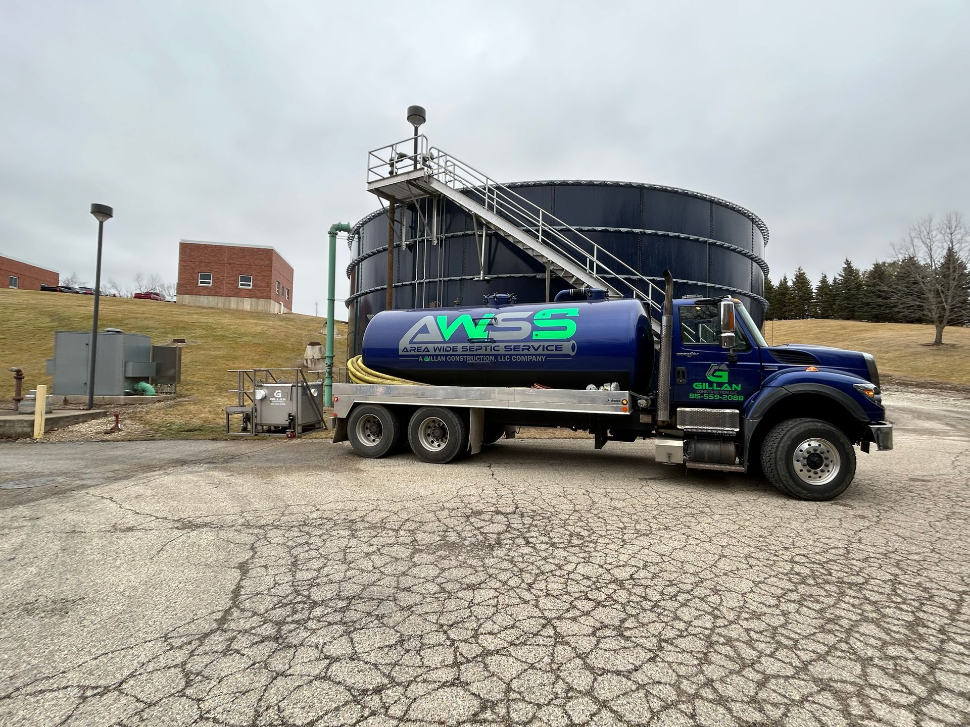 Blue tanker truck with AWSS logo at a wastewater treatment facility, large black tanks in the background.