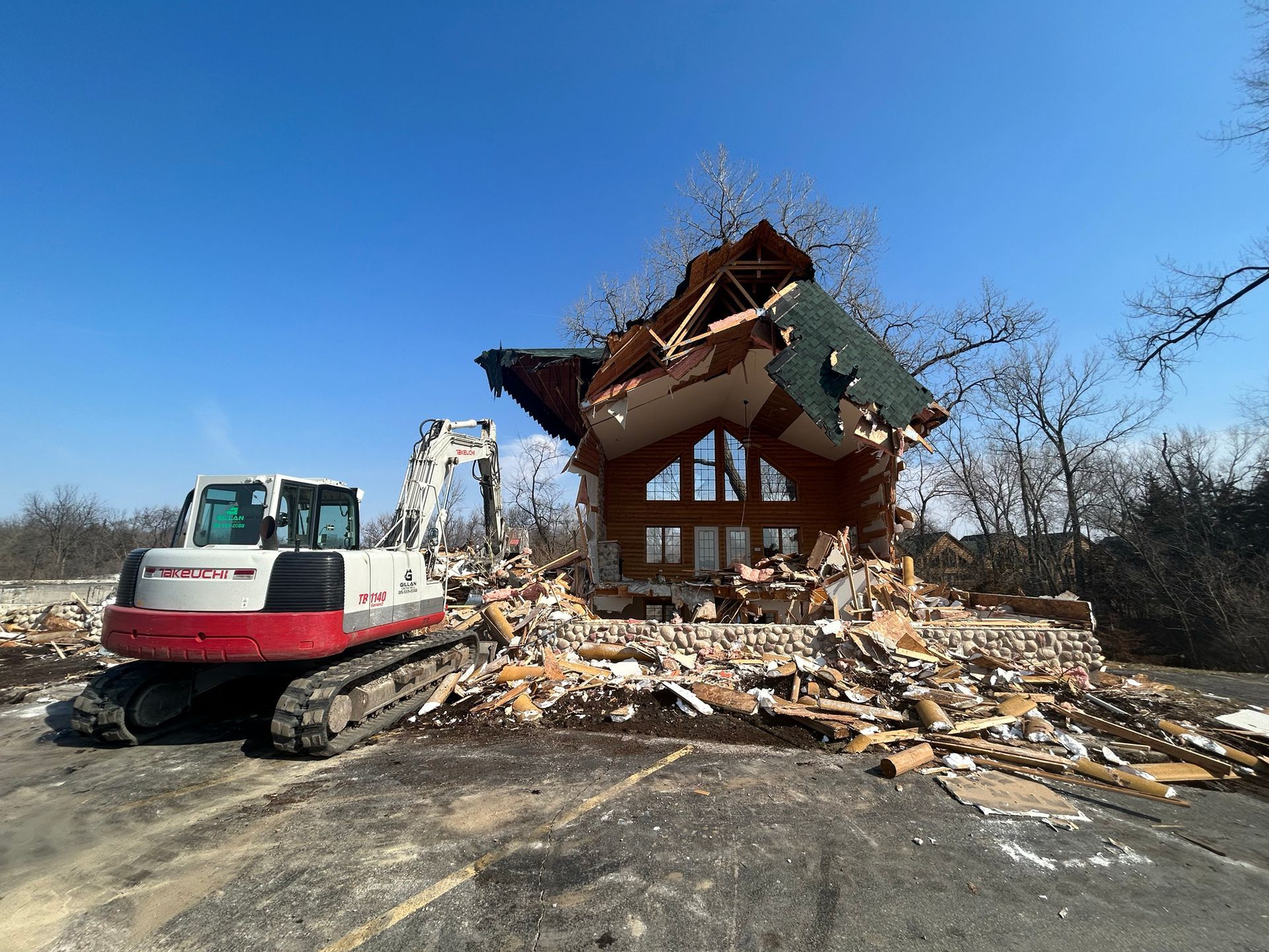 Excavator demolishing a partially-destroyed, multi-story log cabin under a blue sky. Debris scattered around.
