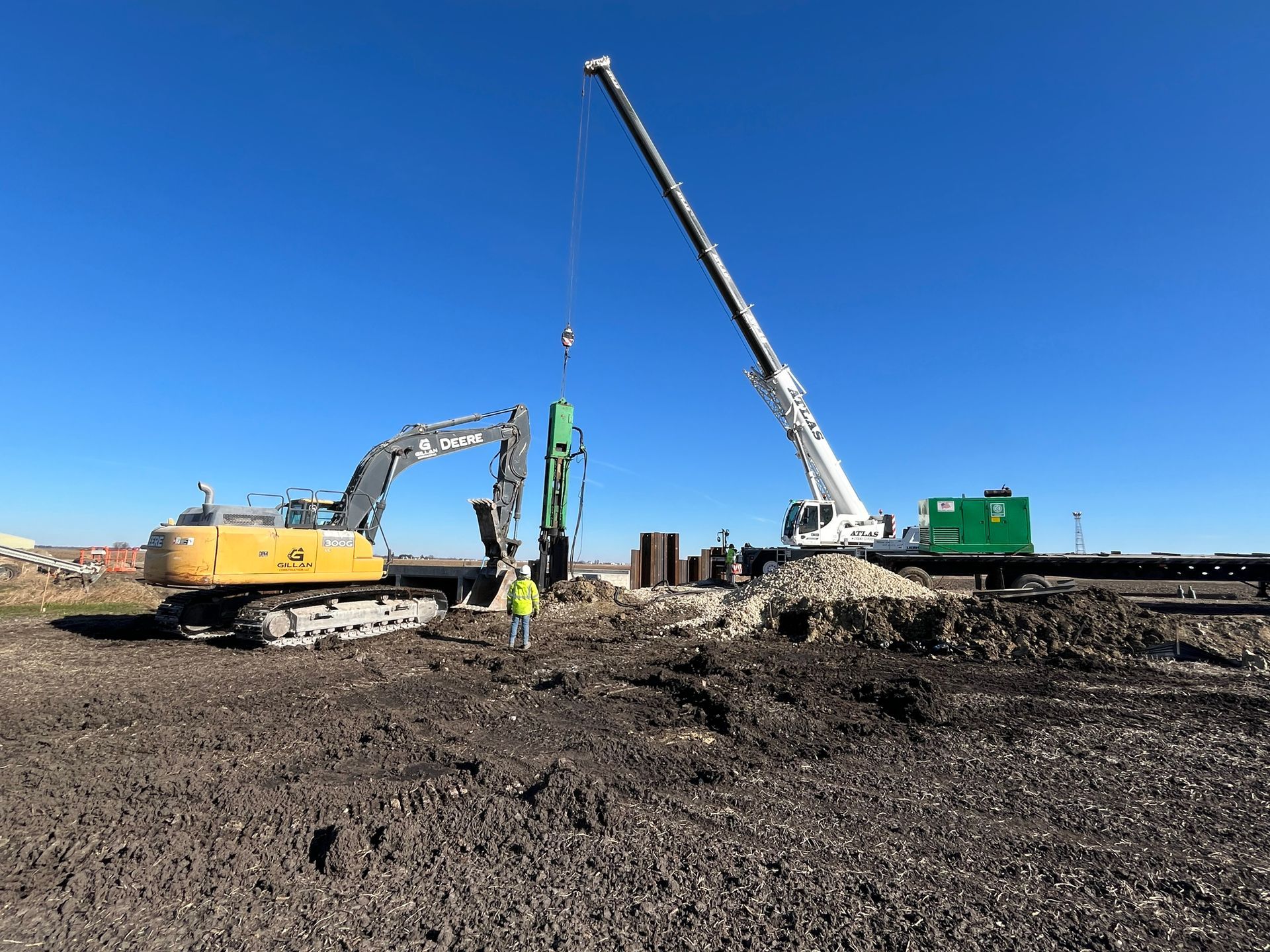 Construction site with excavator, crane, and worker on a brown field under a blue sky.