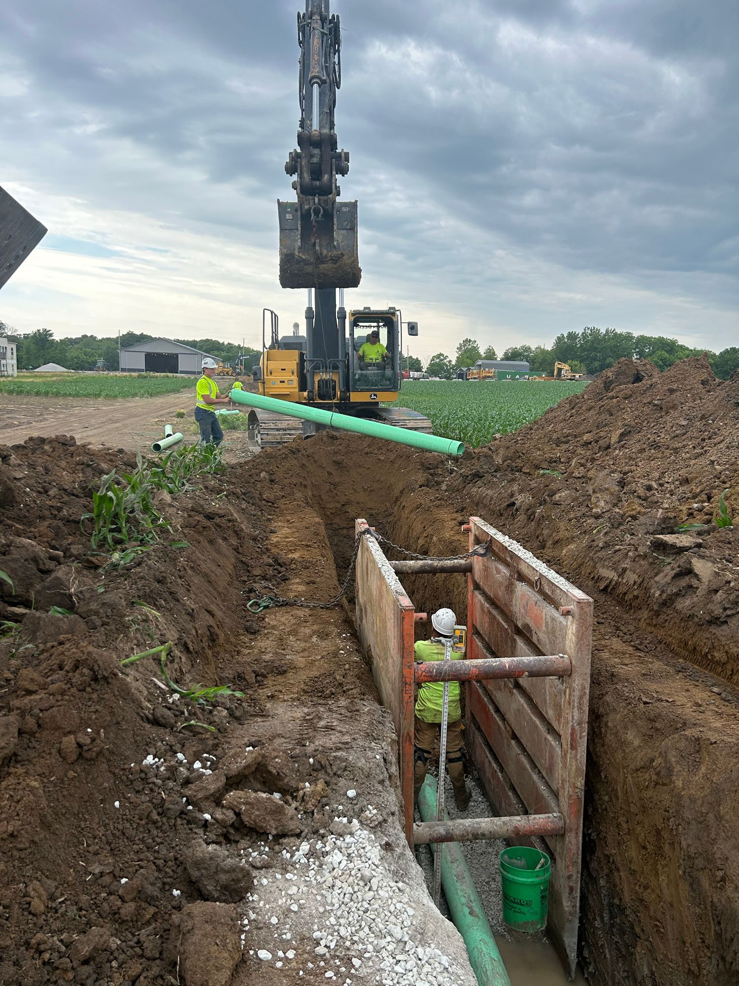 Construction site: excavator and workers installing pipe in trench. Overcast sky, dirt, and equipment visible.