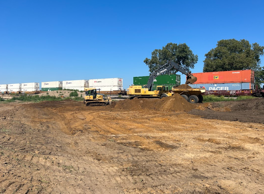 Construction site with bulldozer, excavator, and dump truck; train cars and containers in background.