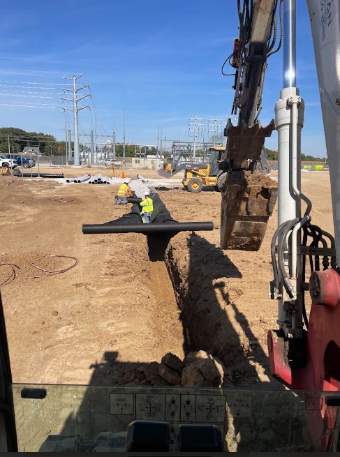 Construction site with excavator digging trench, workers installing pipe, power lines in the background.