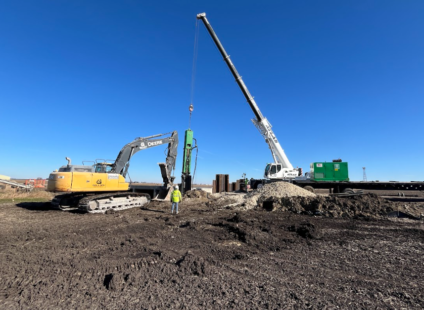 Construction site: Excavator, crane, and worker install a pile under a clear blue sky.