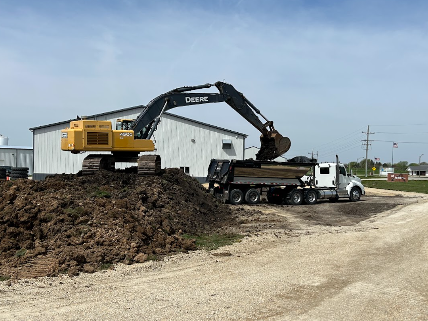 Yellow excavator loading dirt onto a white dump truck near a building on a gravel lot.