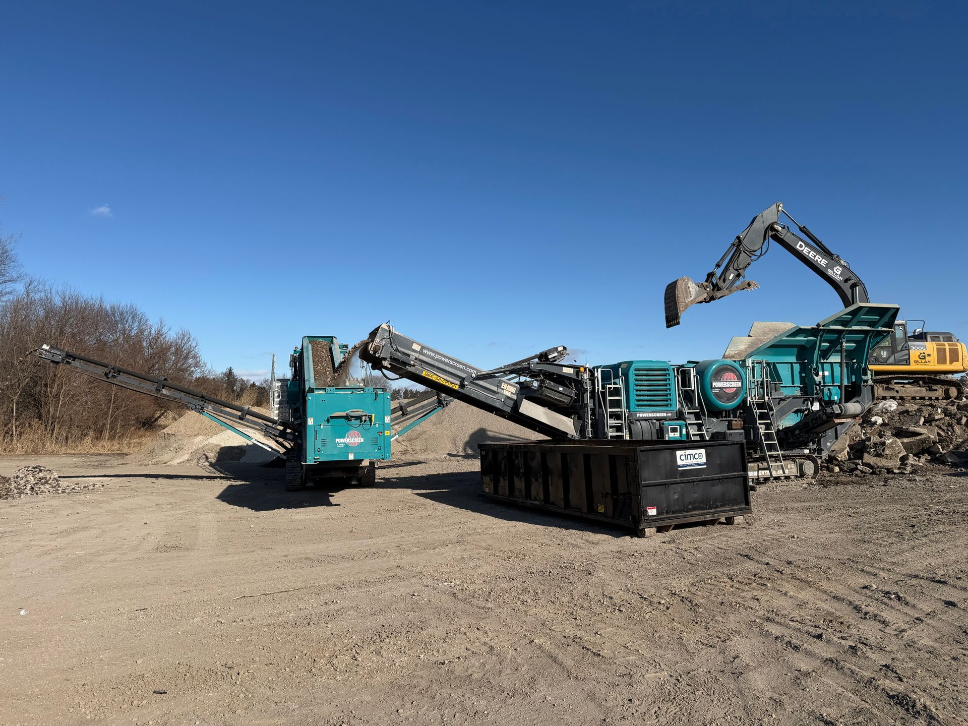 Three teal industrial machines on gravel, processing rubble under a clear blue sky.