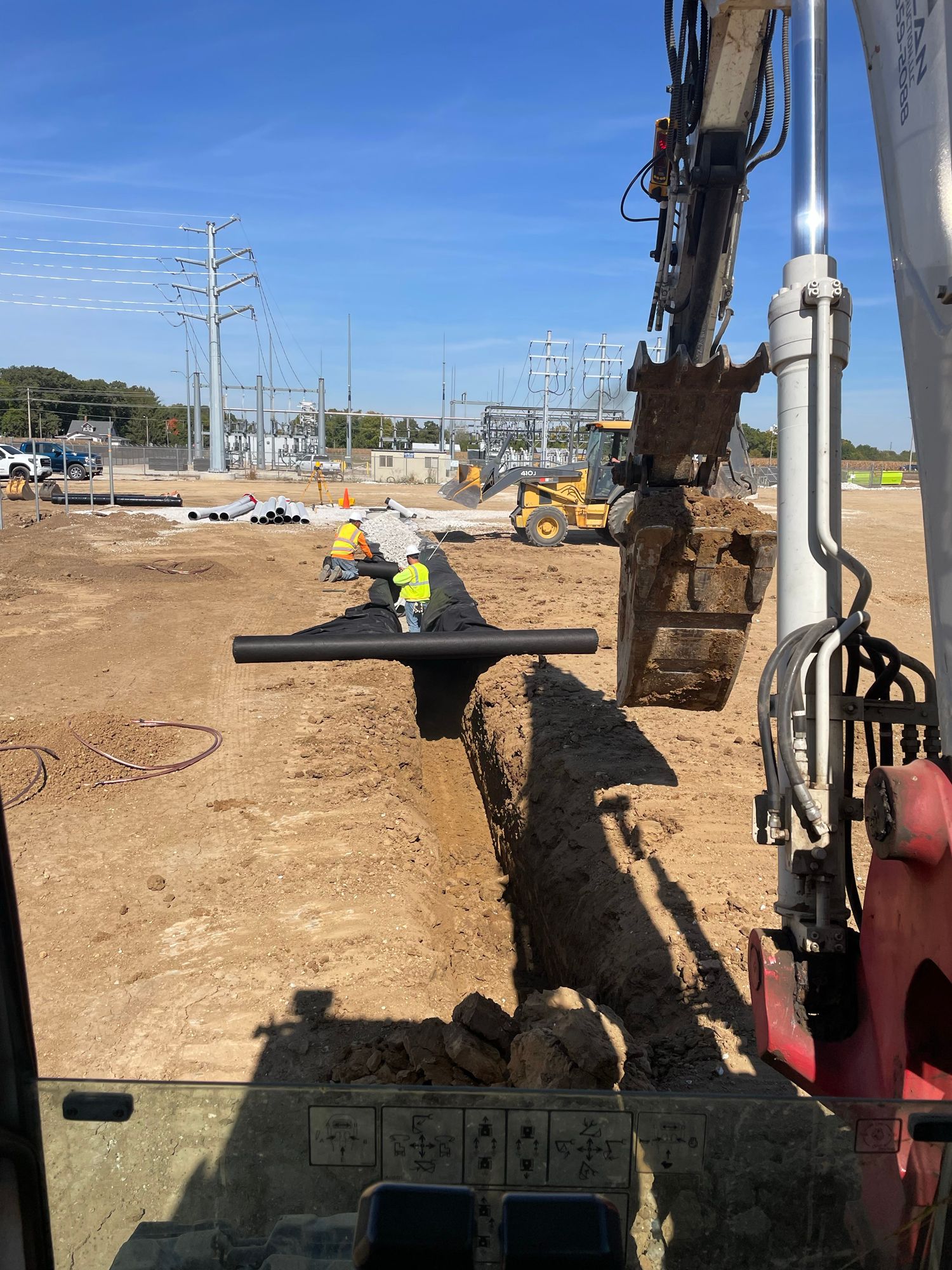 Construction site: excavator placing black pipe in trench; workers in distance.
