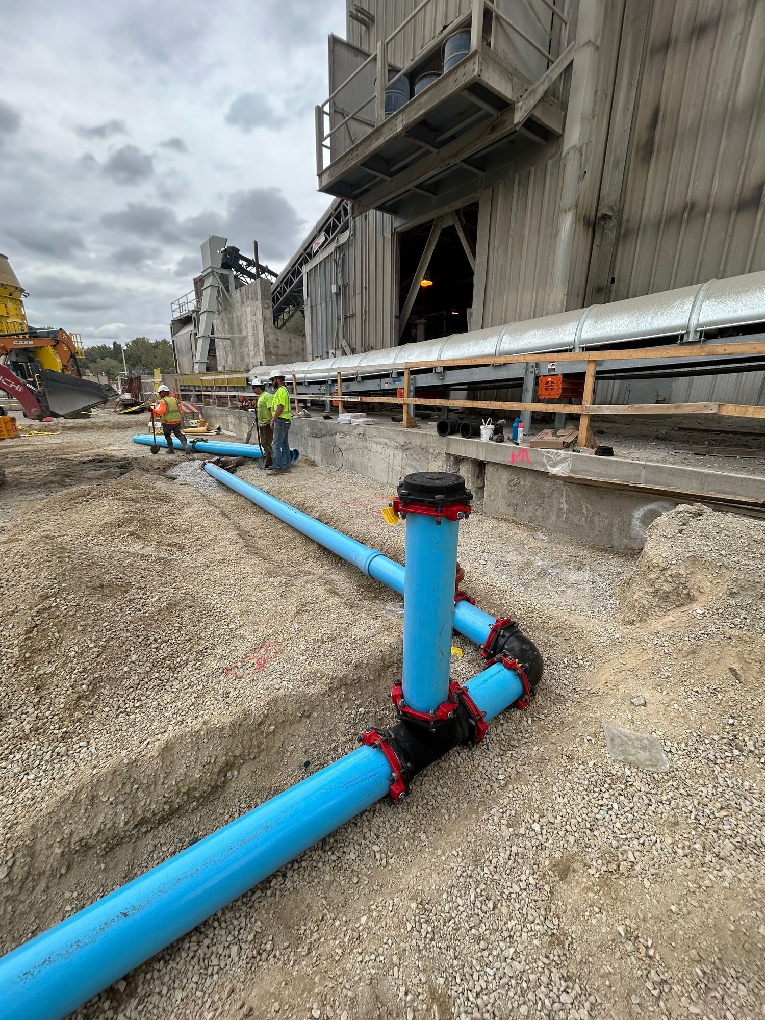 Construction site with blue pipes being installed. Workers in safety vests near a large industrial building.