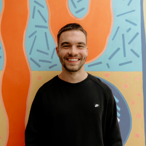Smiling man in a black shirt standing before a colorful abstract mural