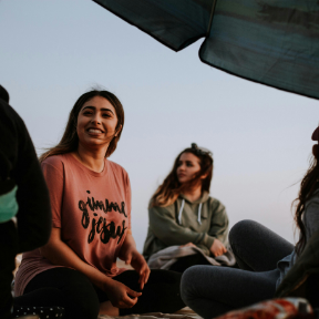 Three friends sitting outdoors under a canopy at sunset, smiling and chatting