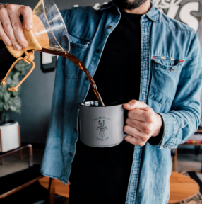 Person pouring coffee into a gray mug in a café, wearing a denim shirt.