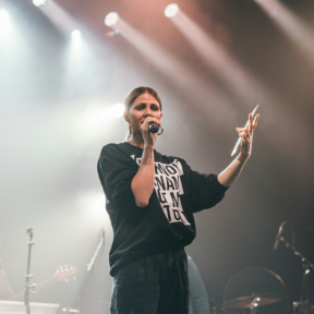 Singer performing onstage with microphone under bright spotlights, wearing a black outfit and gesturing with one hand.