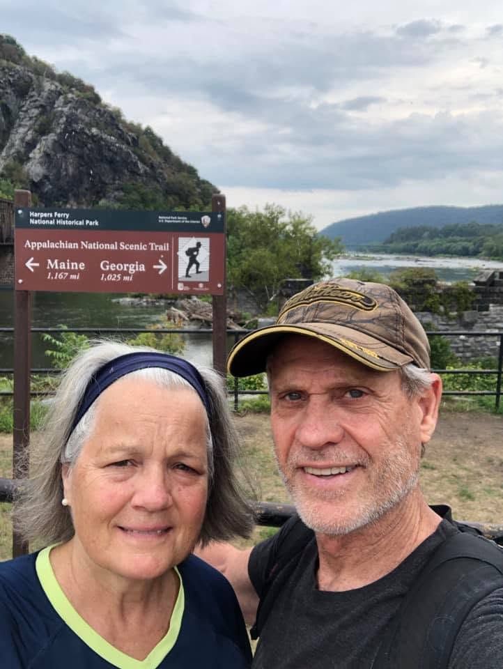Two hikers smiling at a scenic trail sign with mountains and a valley in the background.